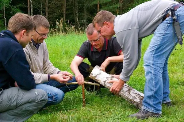 Vier Personen arbeiten gemeinsam an einem Holzstück im Gras beim Überlebenstraining