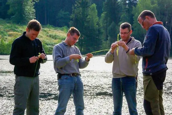 Vier Personen flechten Pflanzenmaterial am Ufer eines Gewässers