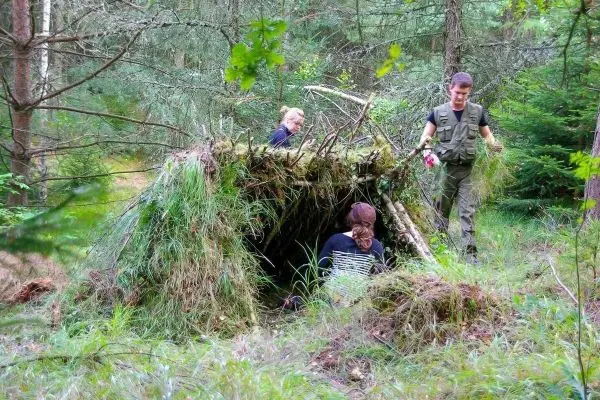 Zwei Personen bauen eine Tarp-Notunterkunft aus Ästen und Gras im Wald