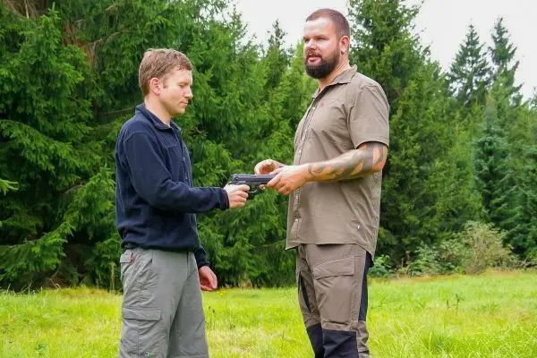 Zwei Personen stehen auf einer Wiese und tauschen eine Handfeuerwaffe aus