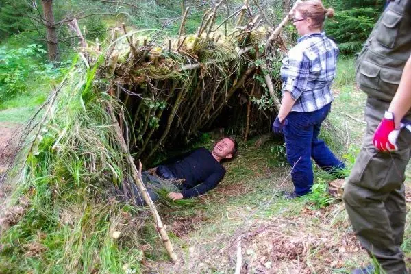 Zwei Personen stehen neben einer selbstgebauten Tarp-Notunterkunft im Wald