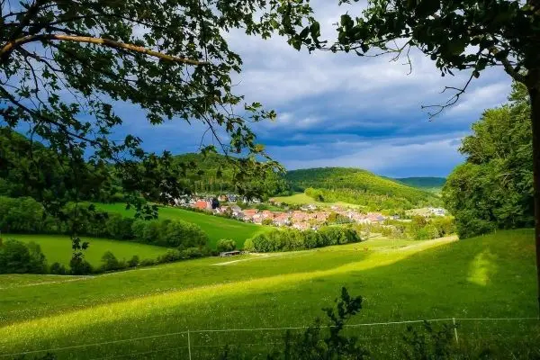 Blick auf eine grüne Wiese mit einem Dorf im Tal und bewaldeten Hügeln im Hintergrund