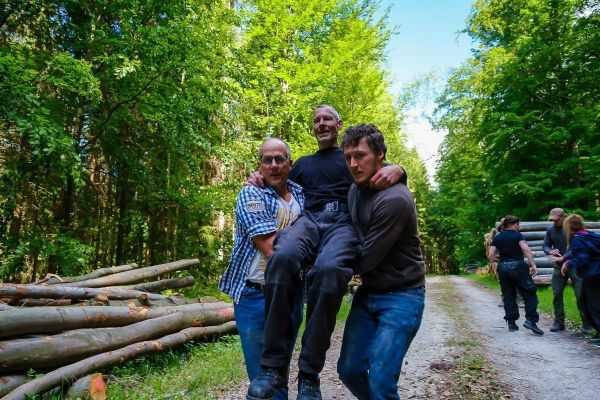 Drei Personen tragen einen Mann auf einem Waldweg zwischen Holzstämmen