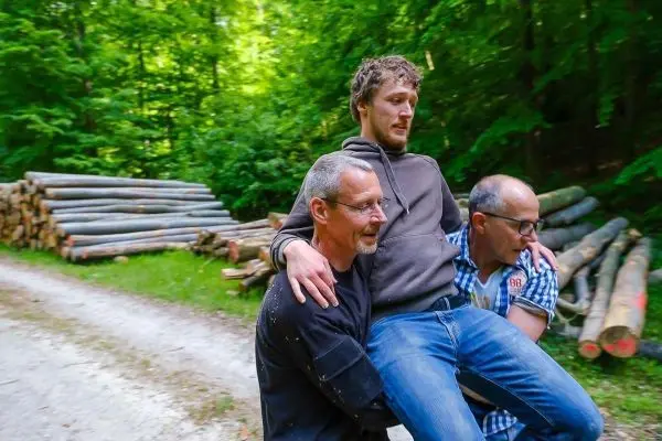Drei Personen tragen einen Mann auf einem Waldweg zwischen Holzstapeln