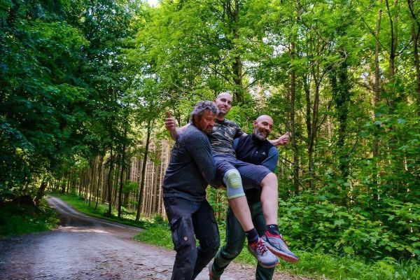 Drei Personen tragen gemeinsam einen Mann auf einem Waldweg