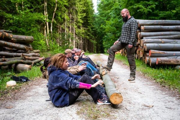 Gruppe sitzt auf dem Boden und beobachtet einen Mann, der auf einem Holzstamm steht