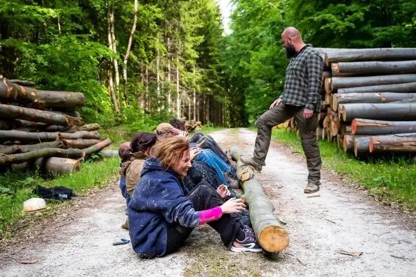 Gruppe sitzt auf einem Baumstamm entlang eines Waldweges, Holzstapel im Hintergrund