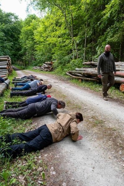Gruppenteilnehmer führen Liegestütze auf einem Waldweg aus, Holzstämme im Hintergrund