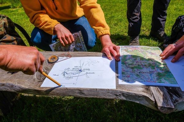 Karte und Kompass liegen auf einer Holzbank, Hände zeigen auf die Zeichnungen