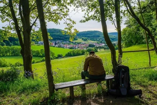 Mann sitzt auf einer Bank und blickt auf eine grüne Landschaft mit einem Dorf