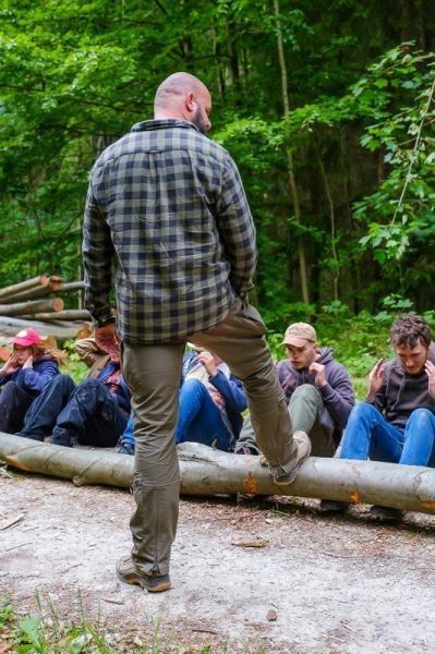 Mann steht auf einem Baumstamm, während mehrere Personen auf dem Boden sitzen