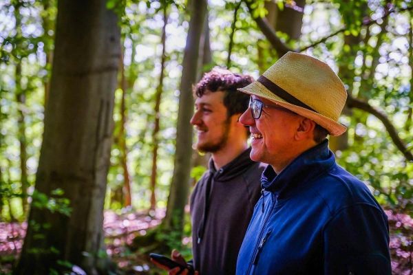 Zwei Männer stehen im Wald und schauen lächelnd in die Ferne