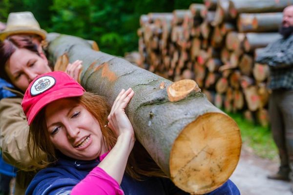 Zwei Personen heben einen Baumstamm, im Hintergrund gestapelte Holzscheite
