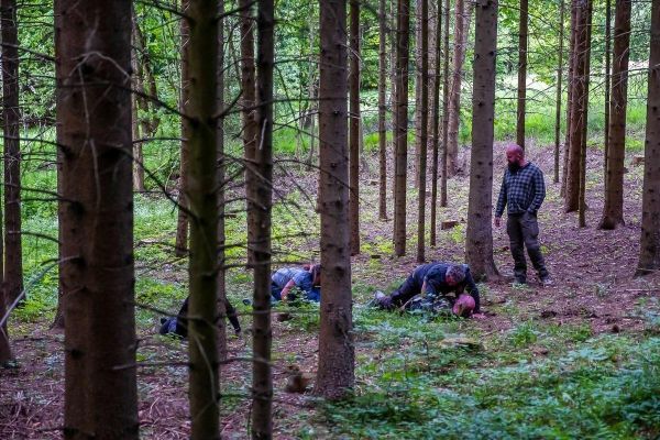 Zwei Personen liegen am Boden, eine steht daneben in einem Wald