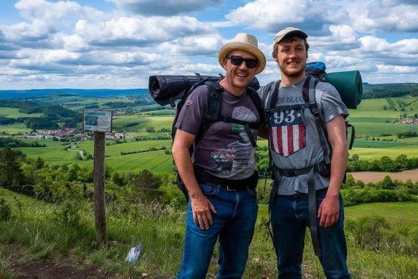 Zwei Wanderer mit Rucksäcken stehen auf einem Hügel mit Aussicht auf die Landschaft