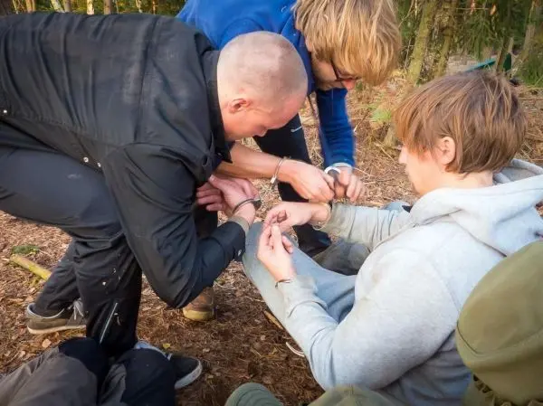 Drei Personen fesseln einen Mann mit Handschellen im Wald
