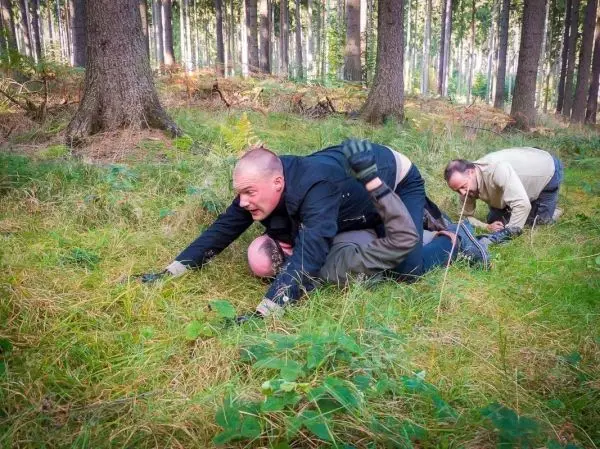 Drei Personen üben Bodenkampf in einem Waldgebiet auf Grasboden