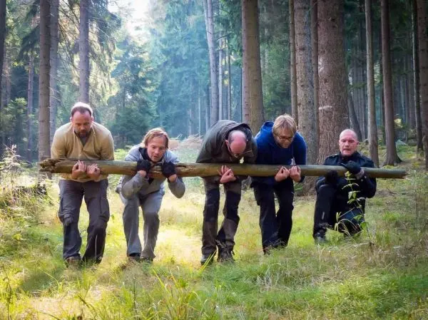 Fünf Personen tragen gemeinsam einen langen Baumstamm durch den Wald