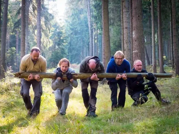 Fünf Personen tragen gemeinsam einen langen Holzstamm durch den Wald