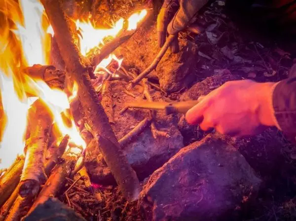 Hand schneidet Holzstücke neben einer kleinen Flamme auf einem Steinhaufen