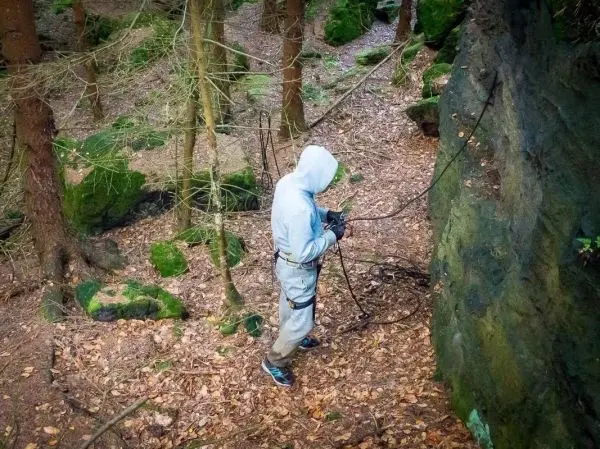 Person hält ein Seil in einem bewaldeten Gebiet mit Felsen und Laub