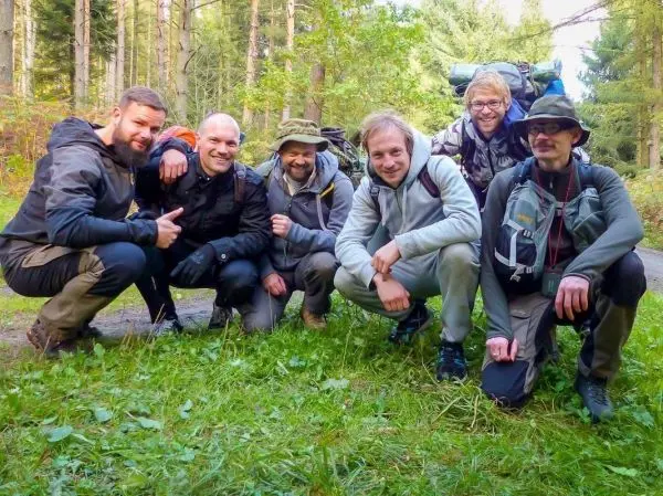 Sechs Personen in Outdoor-Bekleidung posieren lächelnd auf einer Wiese im Wald