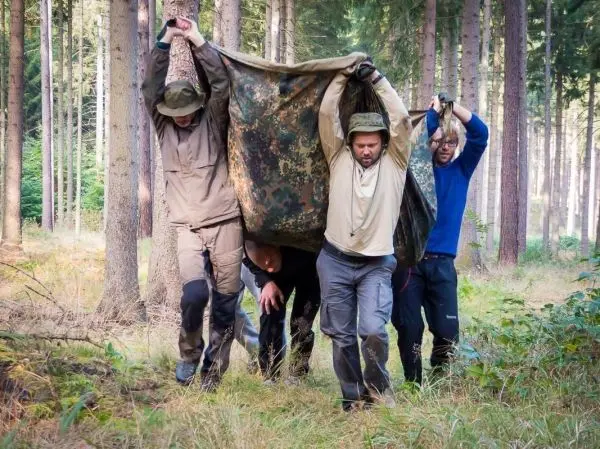 Vier Personen tragen eine improvisierte Trage aus einer Decke durch den Wald