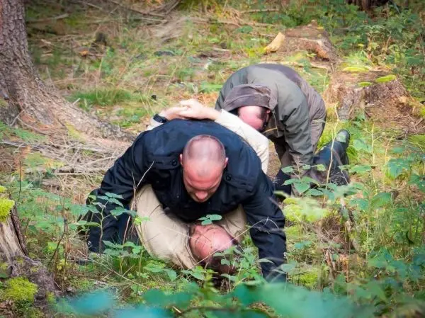 Zwei Personen halten eine dritte Person am Boden in einem Wald fest
