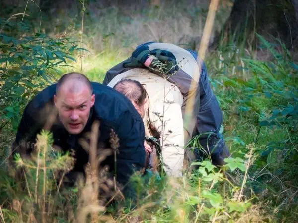 Zwei Personen kriechen durch hohes Gras in einem Waldgebiet