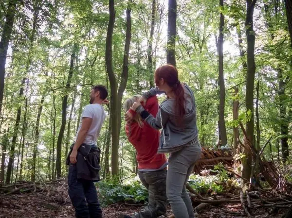 Drei Personen stehen im Wald, eine Person wird von einer anderen festgehalten
