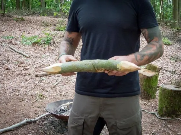 Holzstück mit abgeschälten Enden wird gehalten, im Hintergrund Feuerstelle sichtbar
