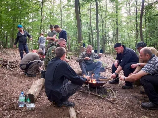 Kochendes Essen über offenem Feuer in einem Waldgebiet, mehrere Personen um den Platz versammelt