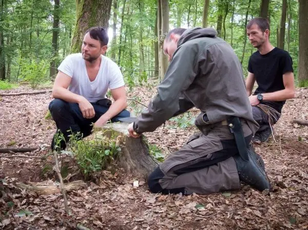 Mann kniet neben Baumstumpf und schnitzt mit einem Messer