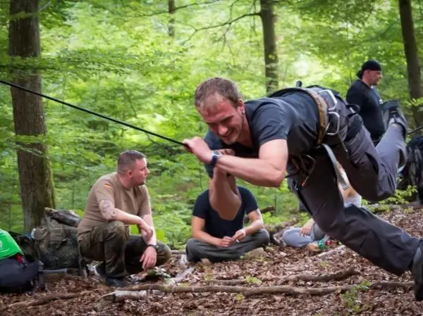 Mann überquert selbstgebaute Seilbrücke im Wald, Zuschauer sitzen im Hintergrund