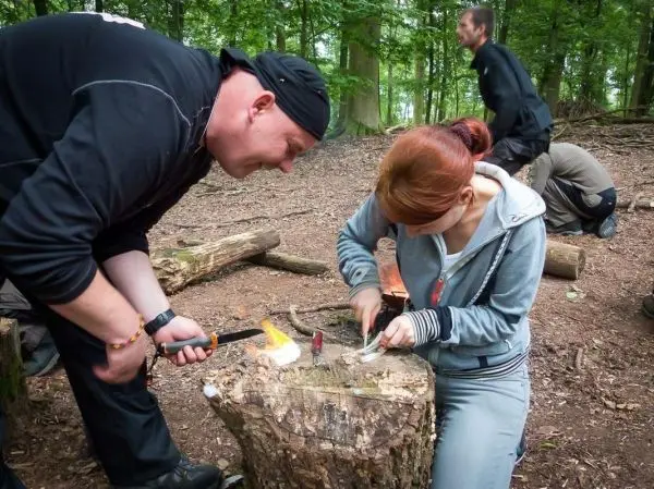 Person schnitzt mit einem Messer an einem Baumstumpf, andere Personen im Hintergrund