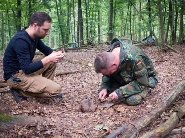 Zwei Personen arbeiten im Wald, einer schnitzt mit einem Messer, der andere beobachtet