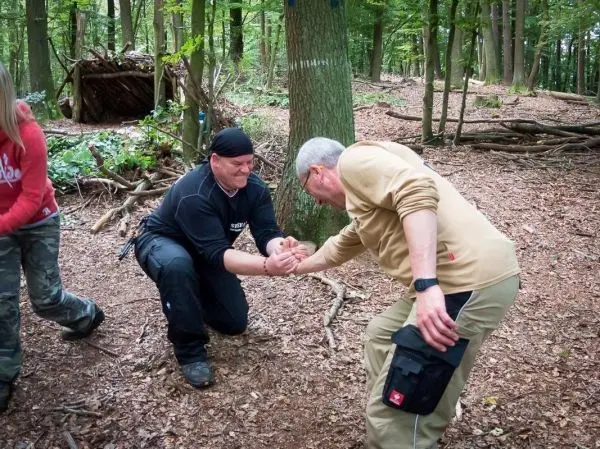 Zwei Personen führen ein Armdrücken im Wald durch, umgeben von Bäumen und Laub