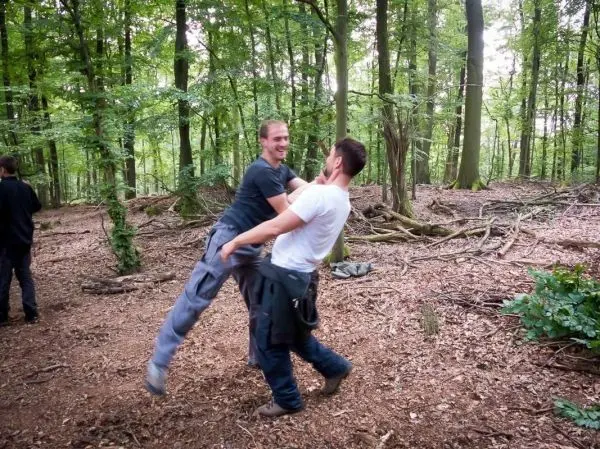 Zwei Personen interagieren spielerisch im Wald, einer hebt den anderen an