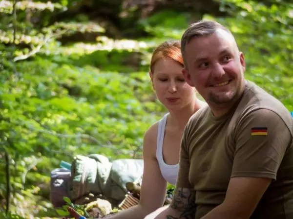 Zwei Personen sitzen im Wald und schauen lächelnd in die Kamera