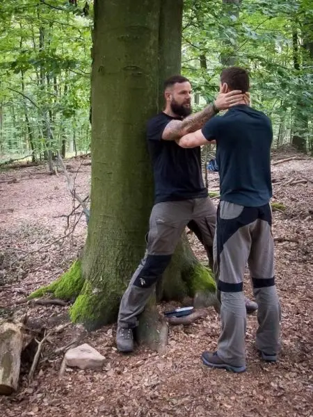 Zwei Personen stehen an einem Baum und üben eine Technik zur Selbstverteidigung