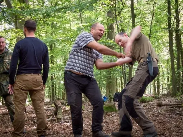 Zwei Personen üben eine körperliche Technik im Wald, umgeben von weiteren Teilnehmern