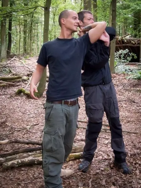 Zwei Personen üben eine Technik im Wald, eine Person hält einen Arm in einer bestimmten Position