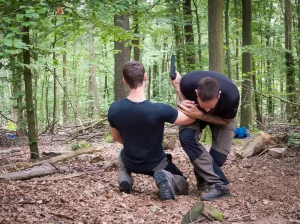 Zwei Personen üben eine Technik zur Kontrolle im Wald, einer kniet und der andere hält seinen Arm