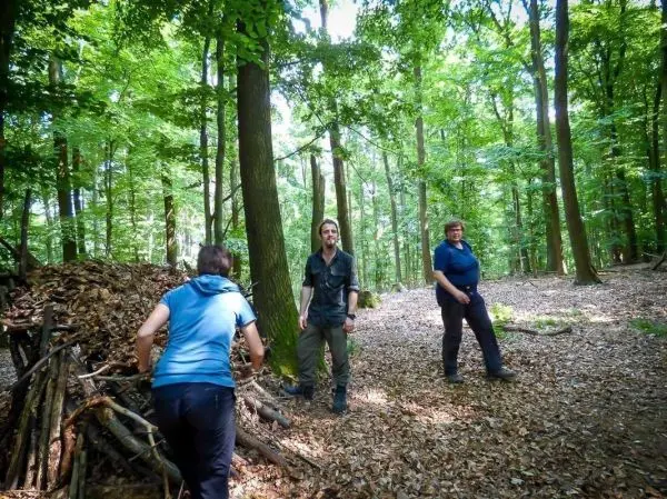 Drei Personen stehen im Wald neben einem Haufen aus Ästen und Laub