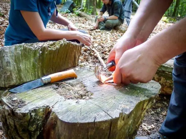 Feuerstahl erzeugt Funken auf Zunder, während ein Messer auf einem Baumstumpf liegt