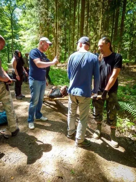 Gruppierung von Personen in einem Wald, eine Person liegt auf einer improvisierten Trage