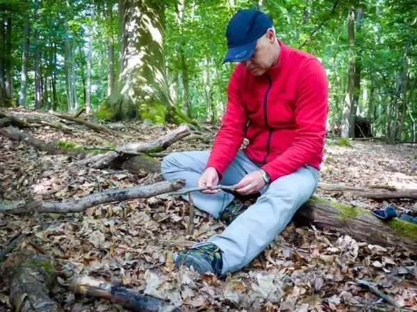 Mann sitzt auf dem Boden und bearbeitet einen Stock mit einem Messer