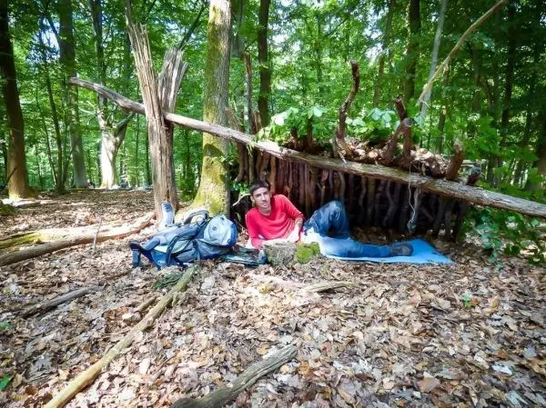 Mann sitzt vor einer selbstgebauten Laubhütte auf einer blauen Unterlage im Wald