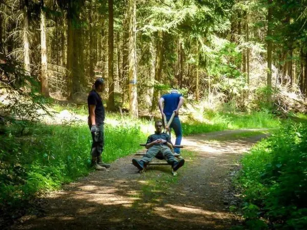 Person liegt auf einer Trage, zwei Personen stehen daneben im Wald