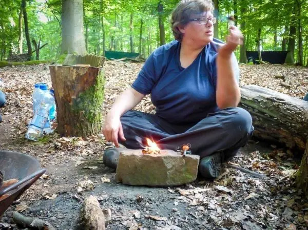 Person sitzt im Wald und hält einen Stock über einer kleinen Flamme